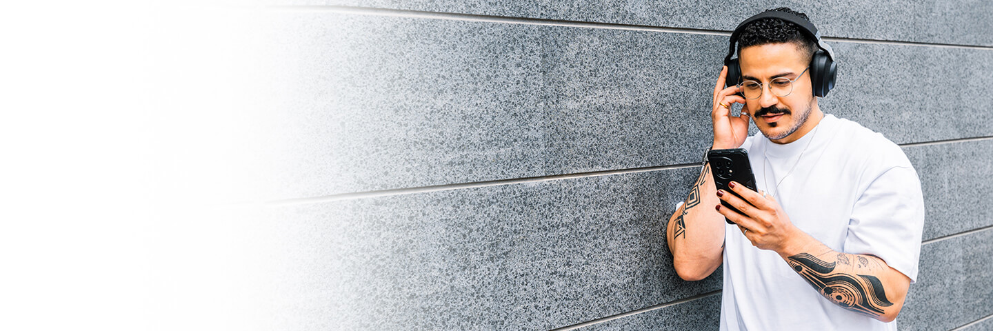 Man listning to headphones and reading his on phone while leaning against a gray stone wall