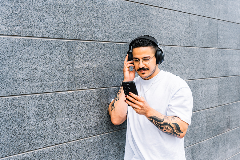 Man listning to headphones and reading his on phone while leaning against a gray stone wall